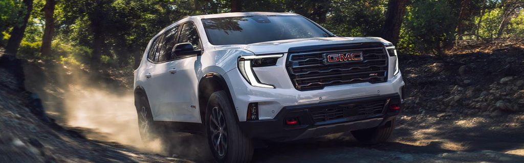White GMC SUV driving off-road through a wooded trail with dust kicking up behind it.